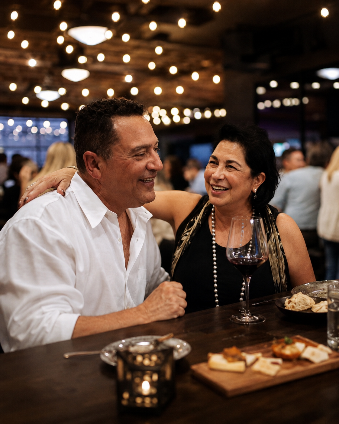A couple laughing over a glass of red wine and a cheese board beneath warm string lights in the winery's tasting room